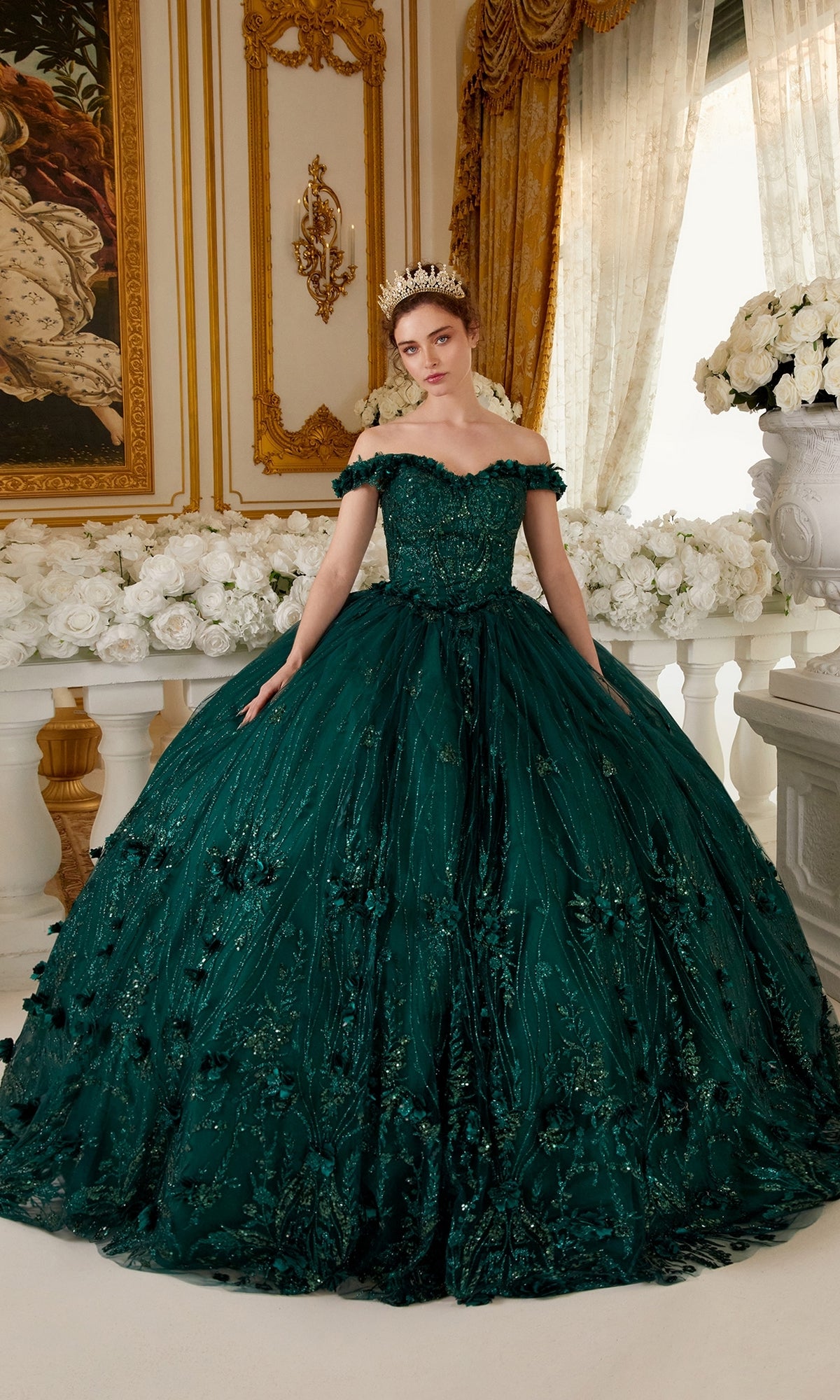 A woman in the Off-the-Shoulder Quince Ball Gown: Ladivine 15704 and a tiara stands in an opulent room with gold accents, white roses, and large windows draped with golden curtains.